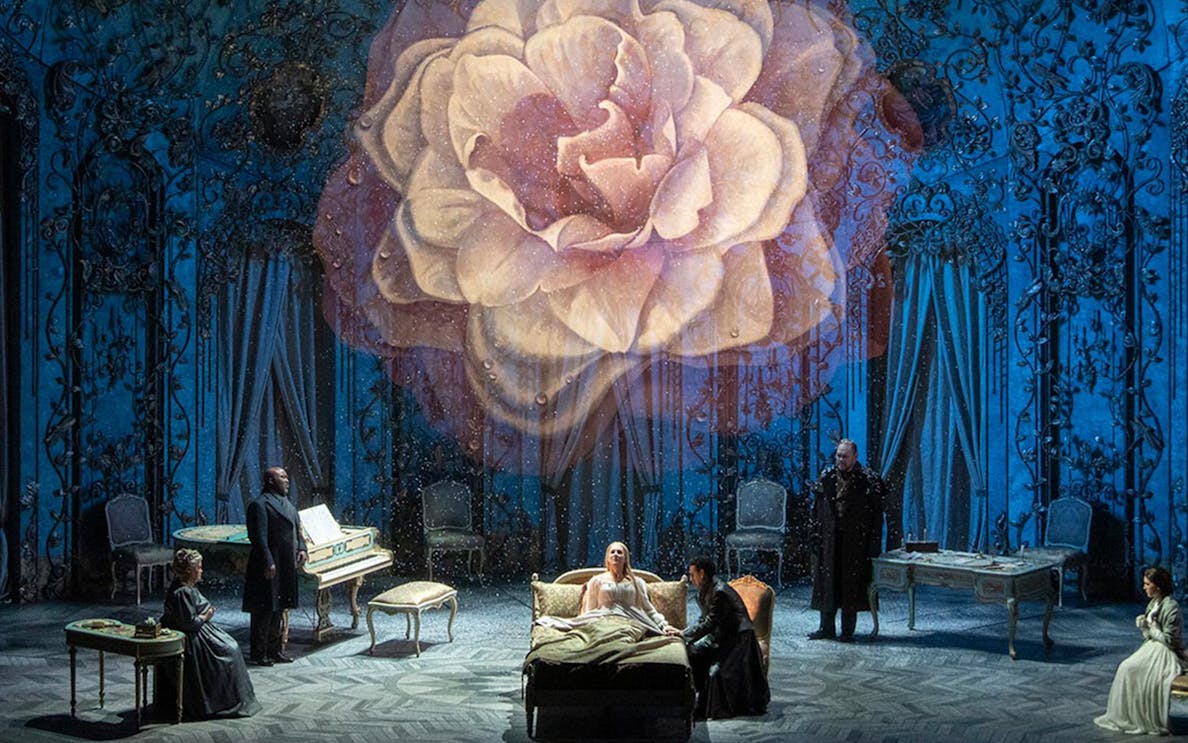 Opera scene from La Traviata with performers on stage and a large flower backdrop.
