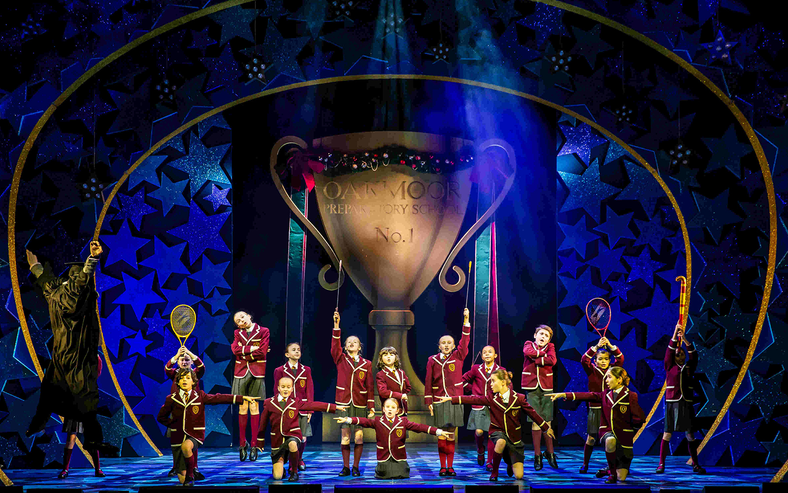 Children in school uniforms perform on stage in Nativity! The Musical, with a large trophy backdrop.