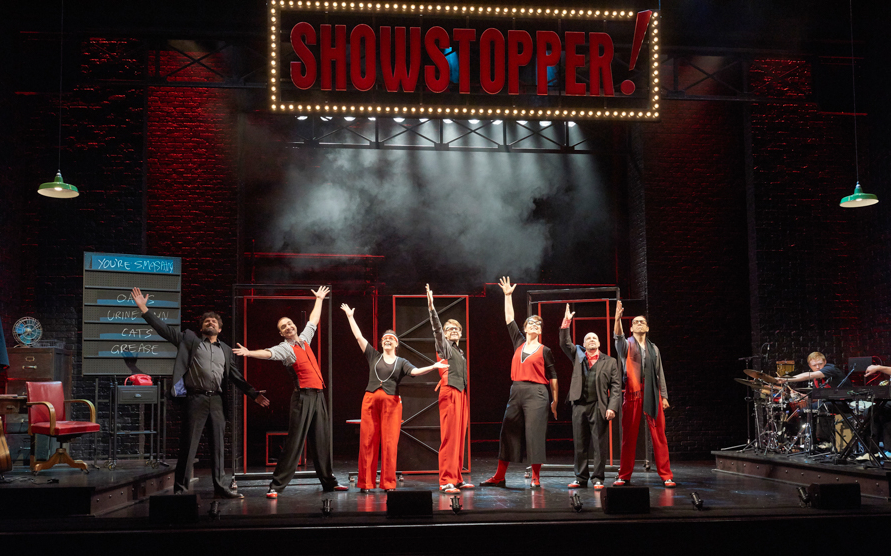 Performers on stage during Showstopper! The Improvised Musical, with arms raised in finale.