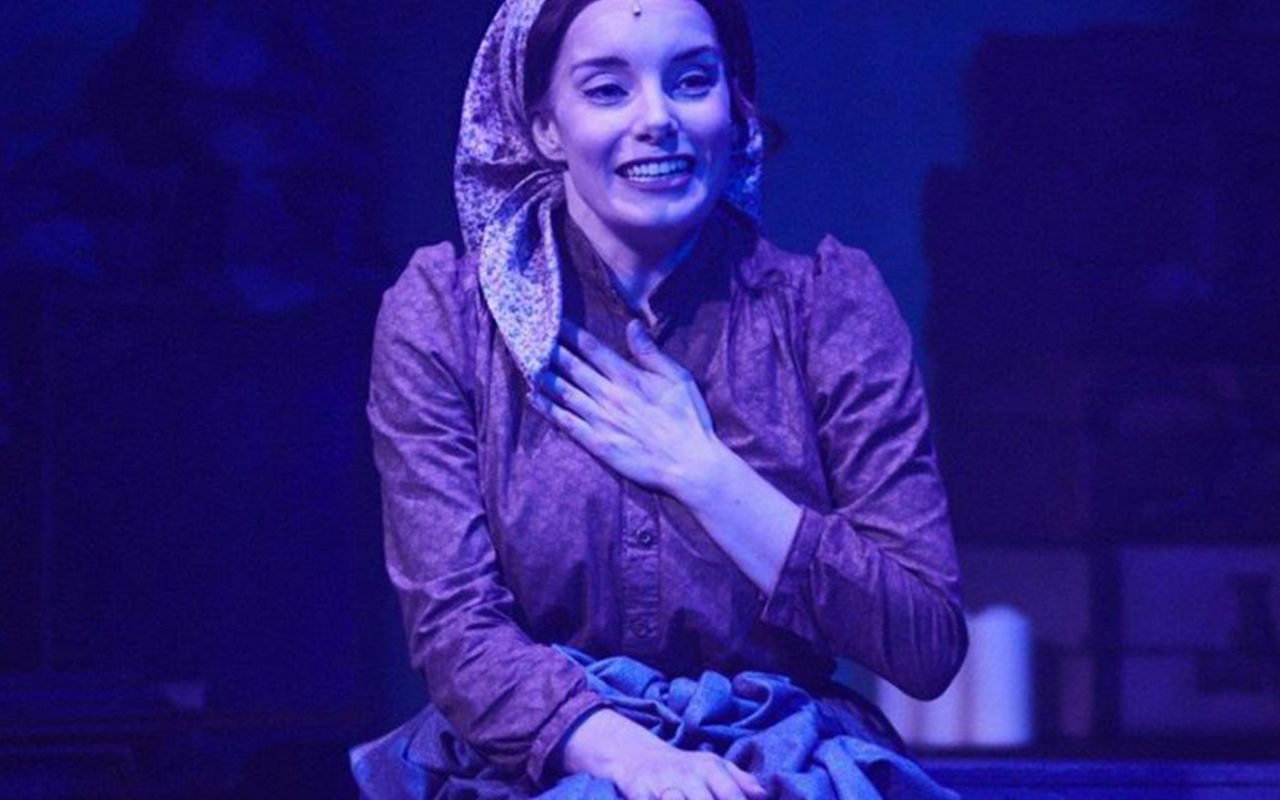 A woman in period costume with a headscarf, hand on chest, performing on stage.