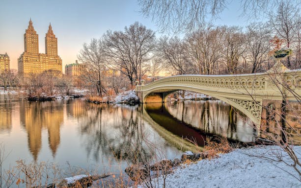 Bow Bridge in Central Park with snow and city skyline in the background.