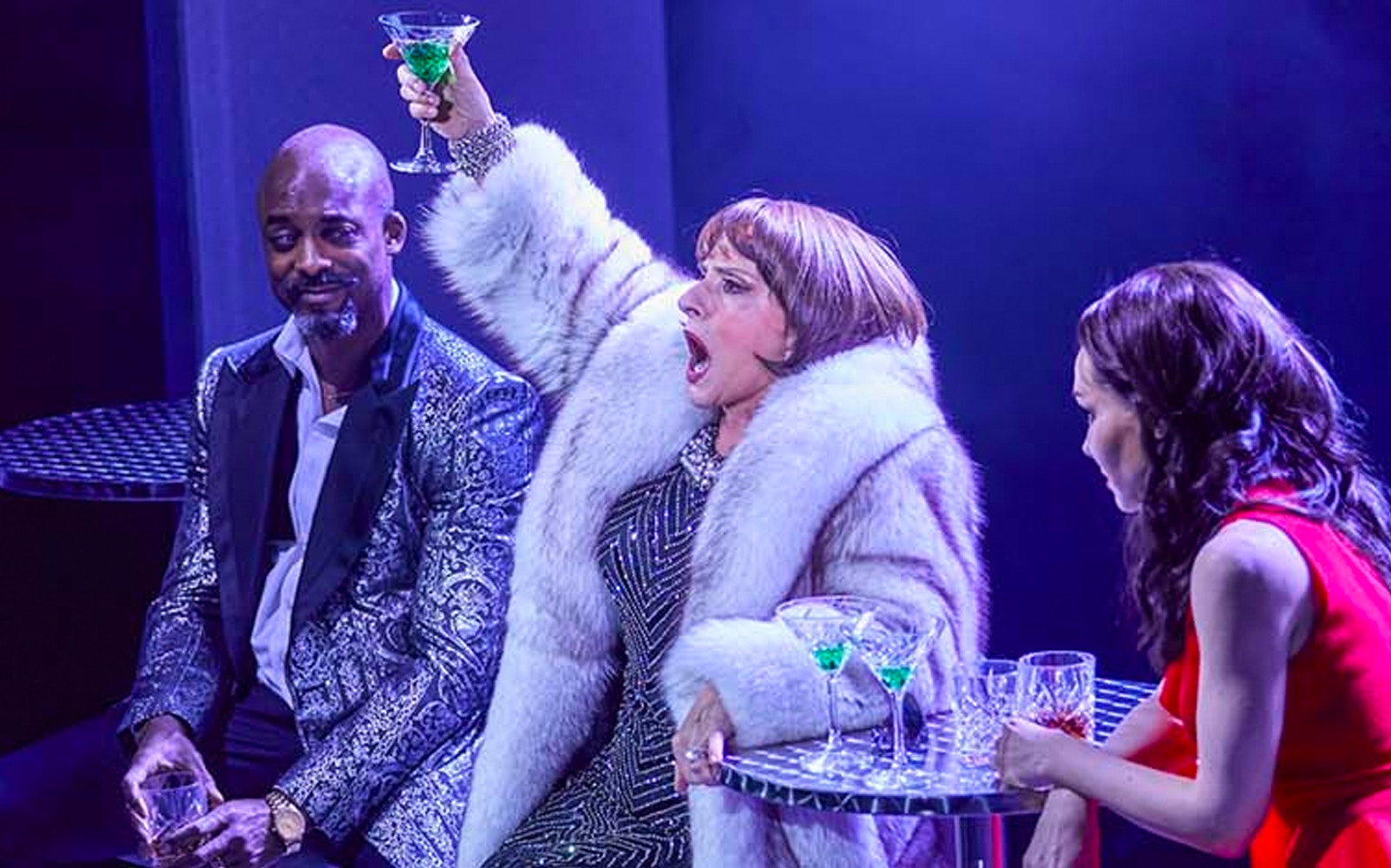 Three people at a table with drinks, one raising a glass in a lively setting.