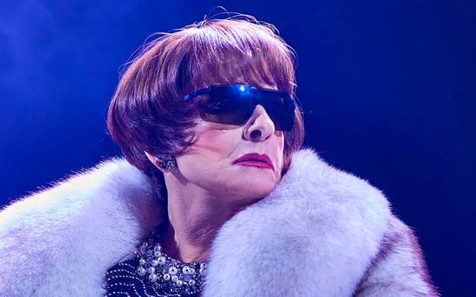 A person wearing sunglasses and a fur coat on stage under blue lighting.