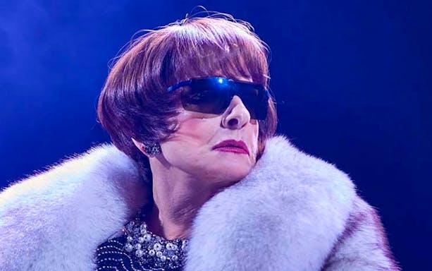 A person wearing sunglasses and a fur coat on stage under blue lighting.