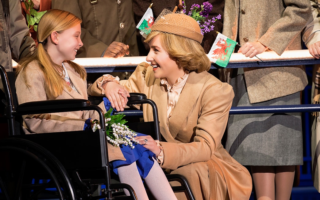 Woman in beige suit smiling at a girl in a wheelchair holding flowers, with Welsh flags in the background.