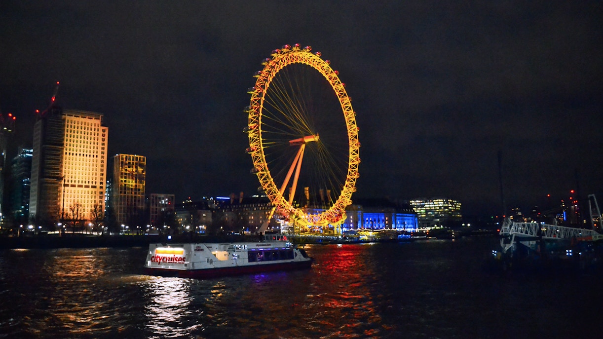 London Eye illuminated at night with a cruise boat on the Thames River.