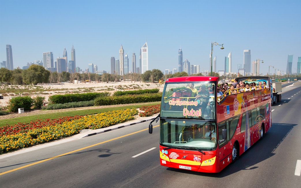 Open-top tour bus on Dubai road with city skyline in background.