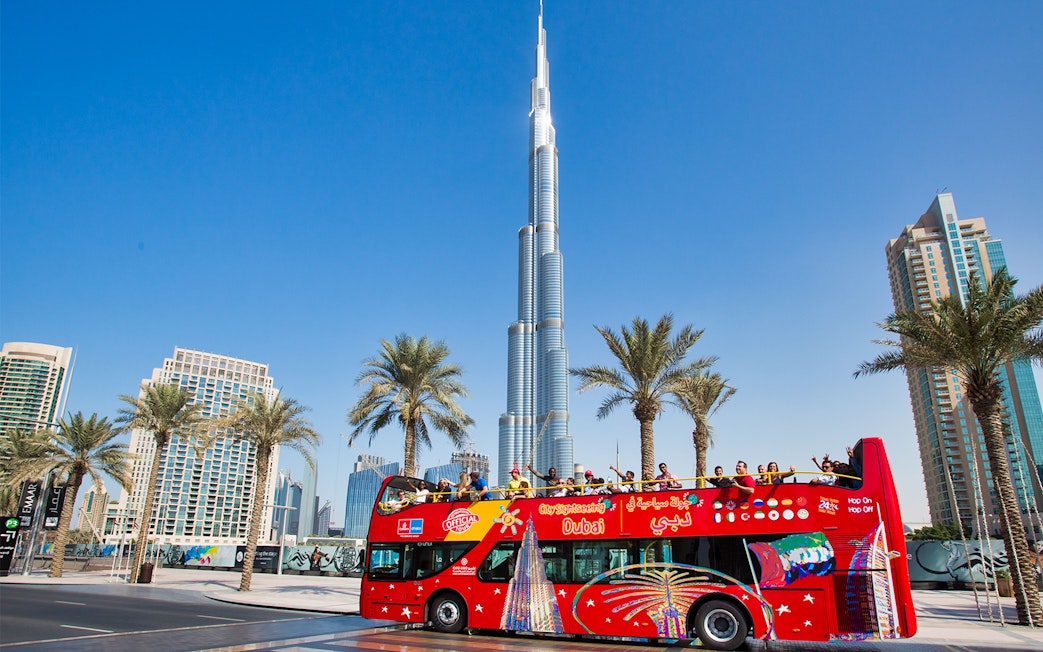 Open-top tour bus near Burj Khalifa, Dubai, with tourists enjoying the view.