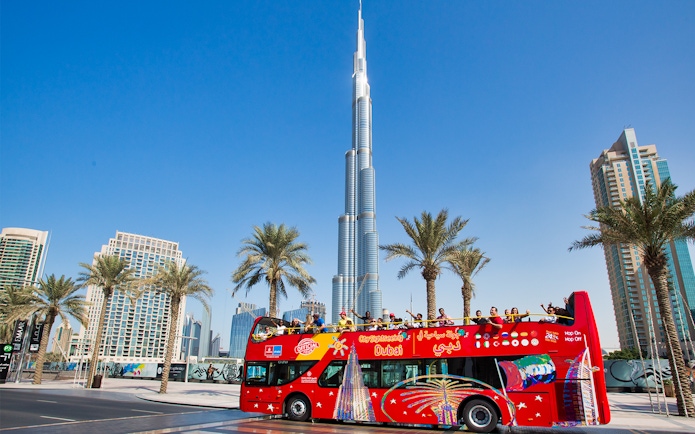 Open-top tour bus near Burj Khalifa, Dubai, with tourists enjoying the view.