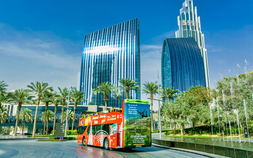 Hop-on-hop-off bus in front of modern skyscrapers and palm trees in Dubai.