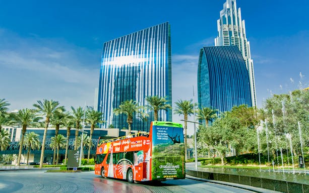 Hop-on-hop-off bus in front of modern skyscrapers and palm trees in Dubai.