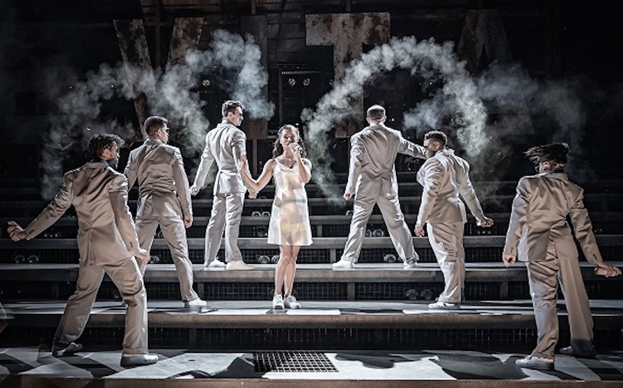 Performers in a theatrical scene from Evita, with dancers in suits on stage.