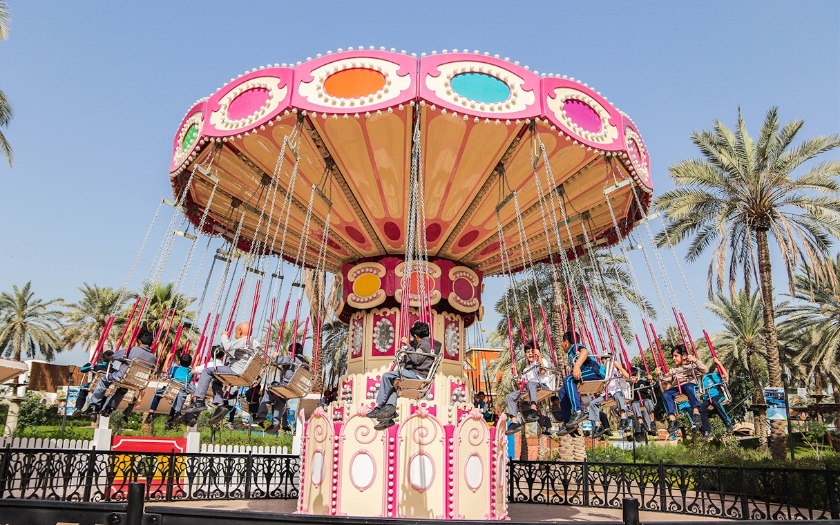 Carousel ride at Al Montazah Water Park with people enjoying swings under palm trees.