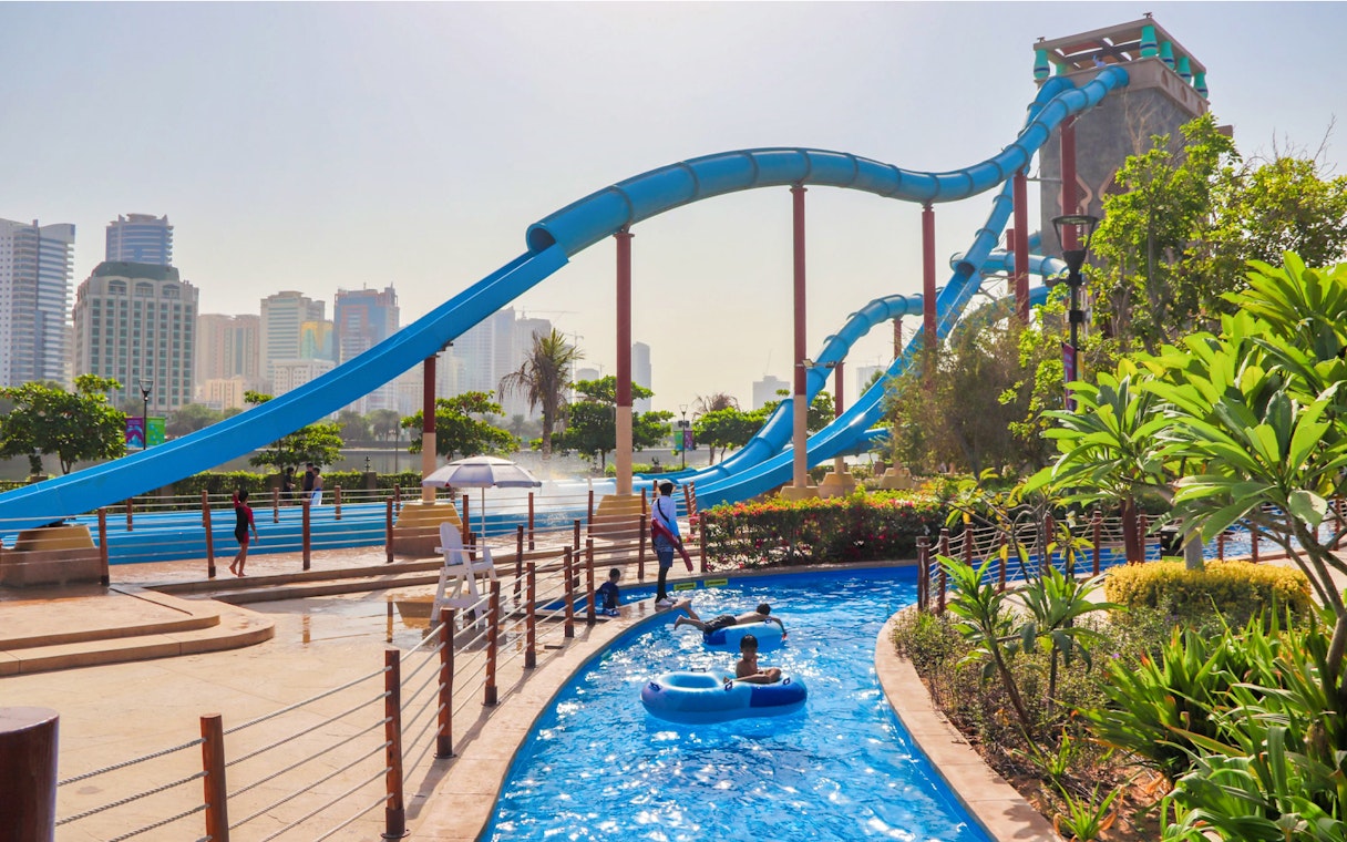 Water slides and lazy river at Al Montazah Water Park, Sharjah, with city skyline in background.
