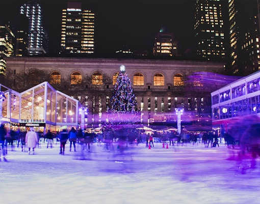 Ice skaters at Rockefeller Center with a lit Christmas tree and city skyline at night.