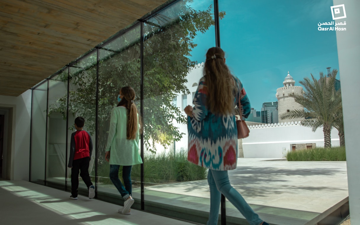 Visitors walking through glass corridor with view of Qasr Al Hosn tower in Abu Dhabi.