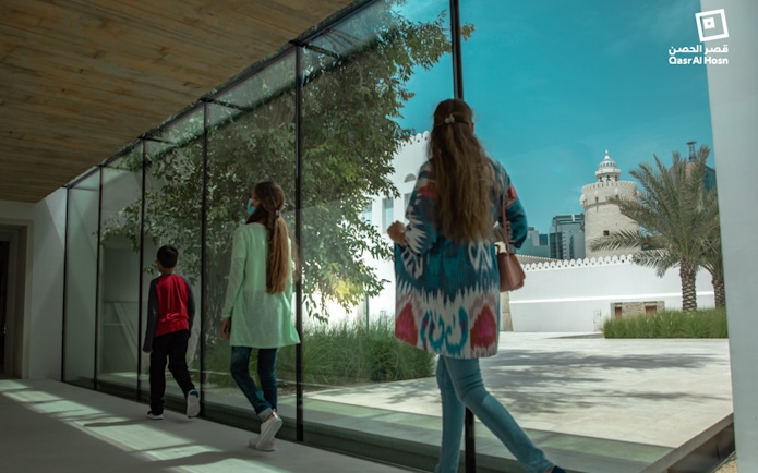 Visitors walking through glass corridor with view of Qasr Al Hosn tower in Abu Dhabi.