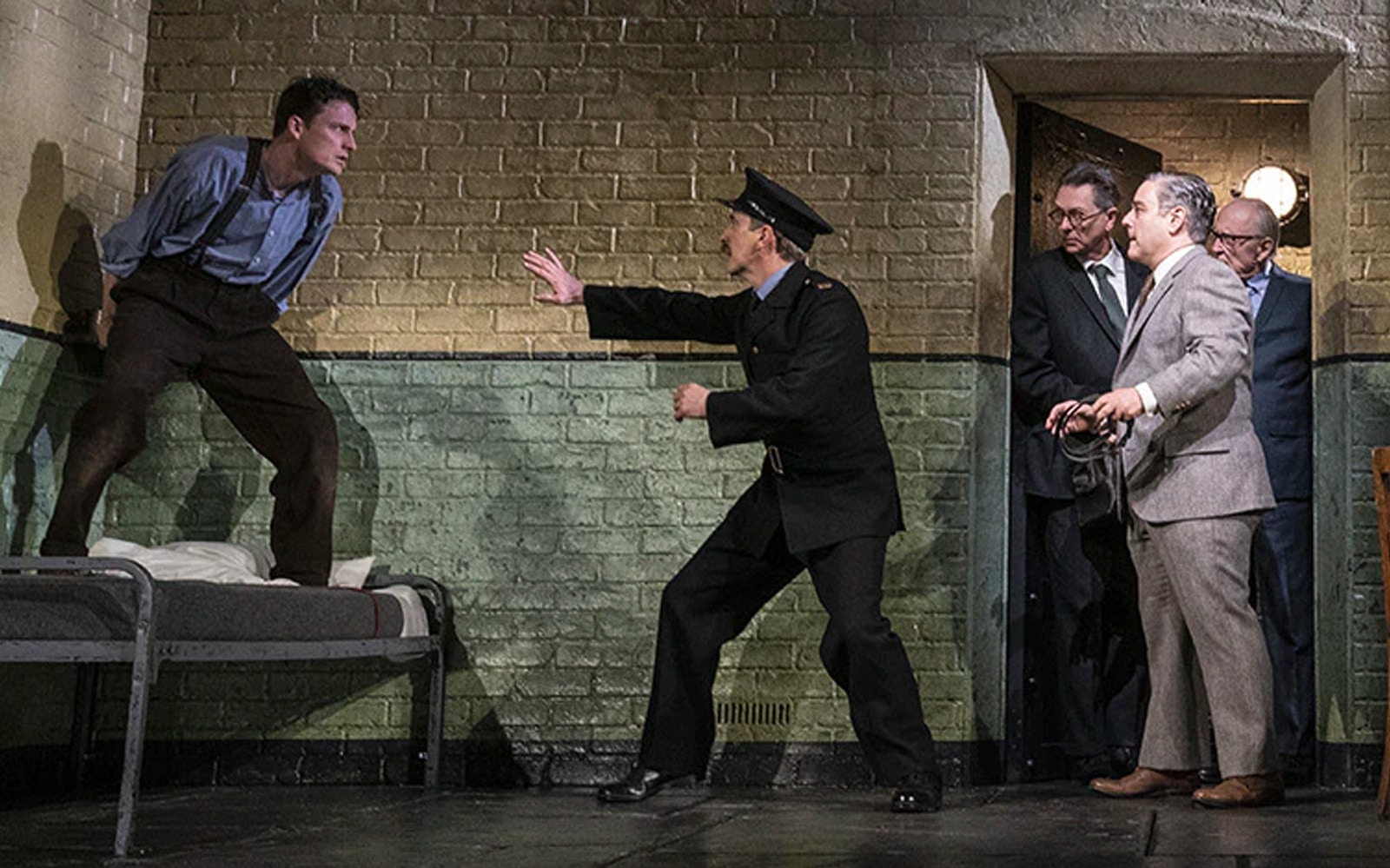Man in uniform reaching towards another man on a bed, with three men observing in a dimly lit room.