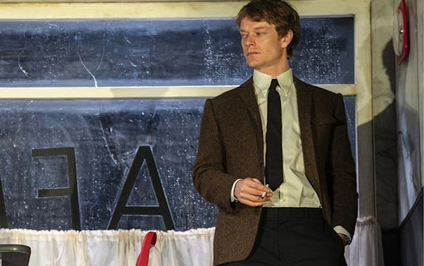 Actor in a scene from the play Hangmen, standing in front of a window with letters visible.