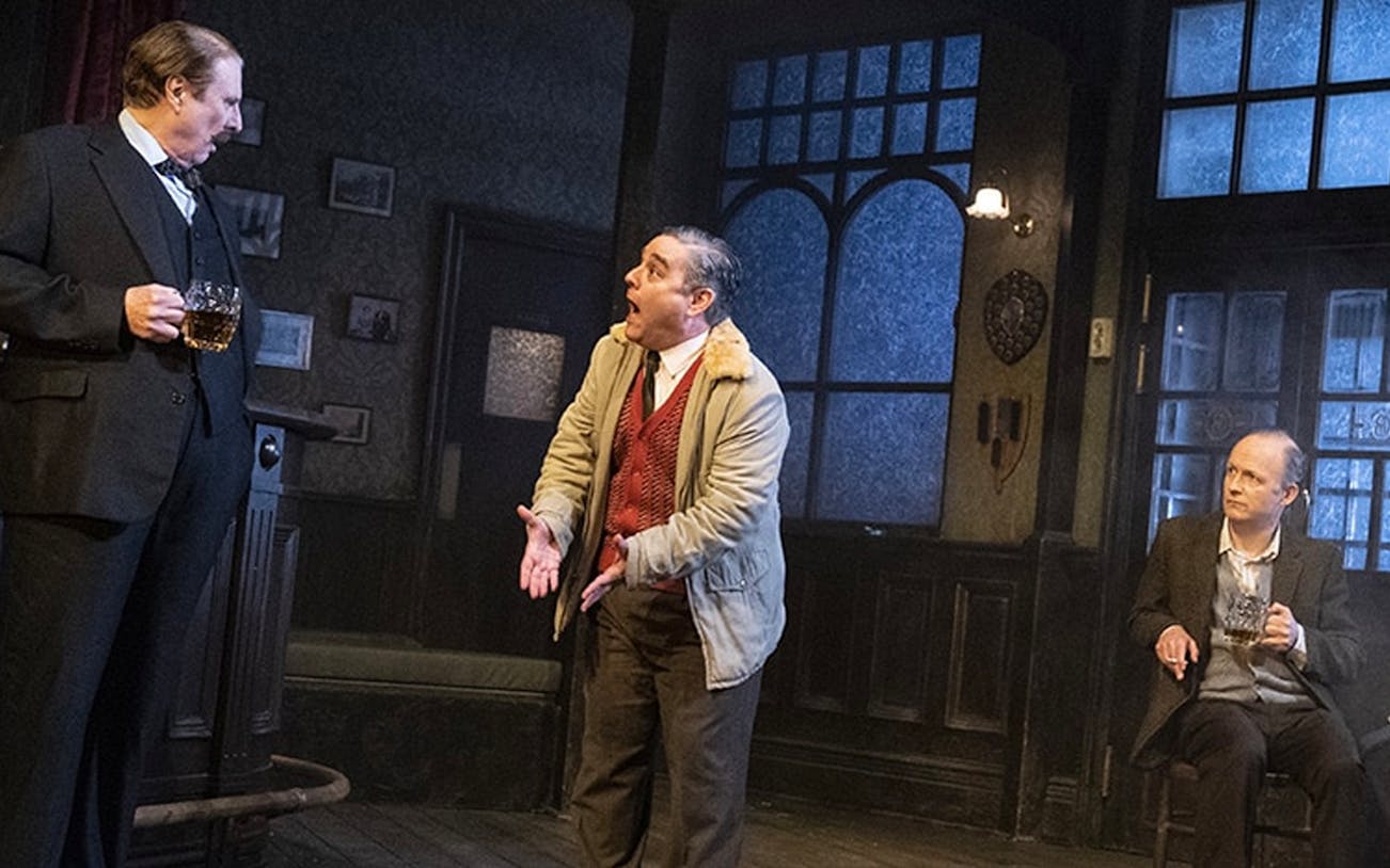 Three actors perform a scene in a dimly lit pub setting from the play "Hangmen.