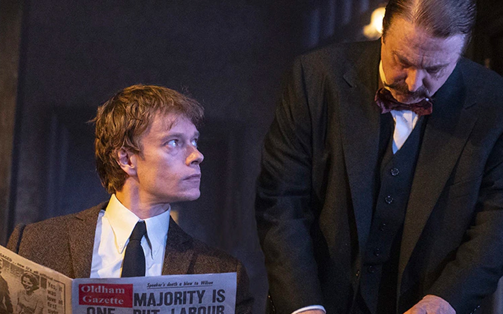 Two men in a dimly lit room, one reading a newspaper titled "Oldham Gazette.