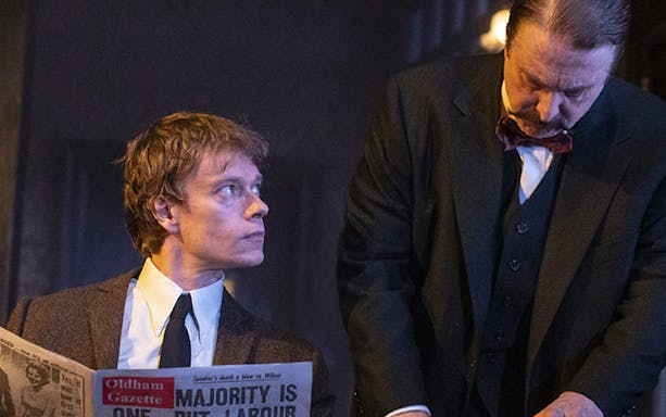 Two men in a dimly lit room, one reading a newspaper titled "Oldham Gazette.