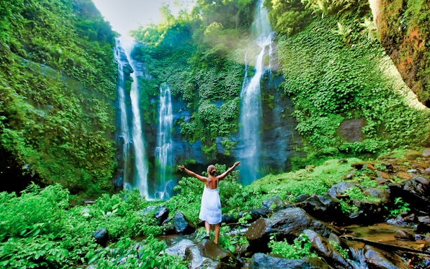 Person standing in front of lush green Bali waterfall, arms outstretched.