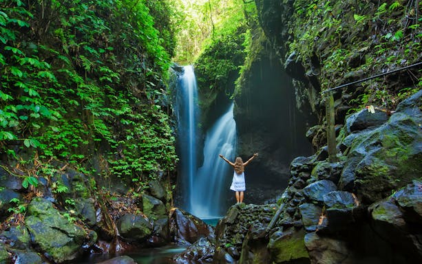 Person standing near a waterfall surrounded by lush greenery in Bali.