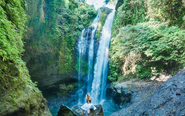 Waterfall surrounded by lush greenery in Bali with a person sitting on a rock.