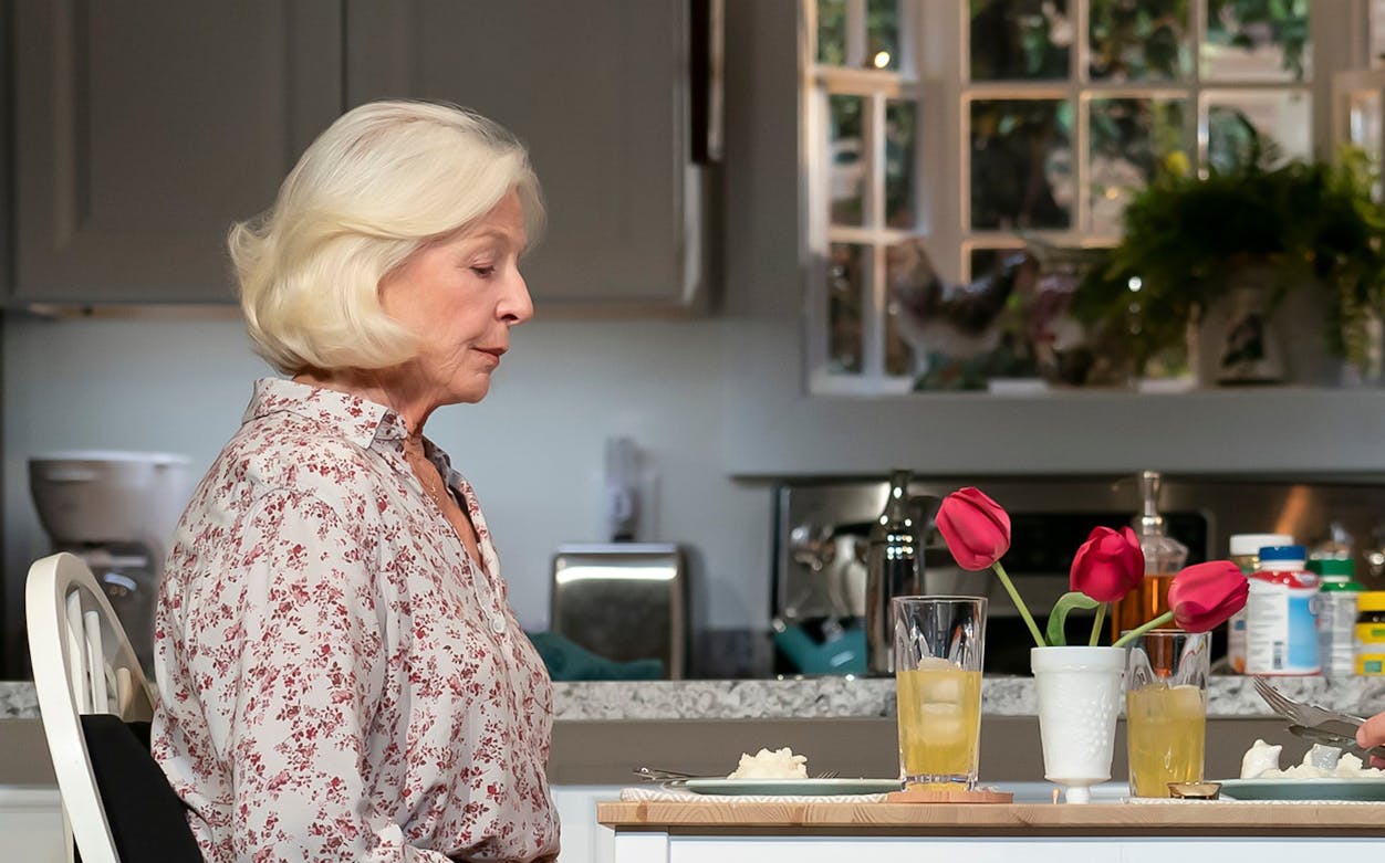 Elderly woman sitting at a kitchen table with drinks and flowers, related to Grand Horizons.