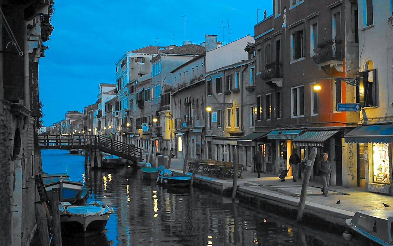 Venice canal at dusk with illuminated buildings and a wooden bridge.