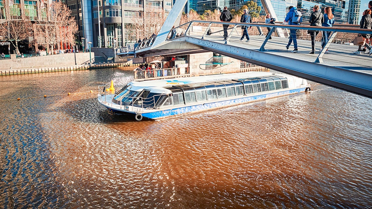River cruise boat passing under a pedestrian bridge in a city docklands area.