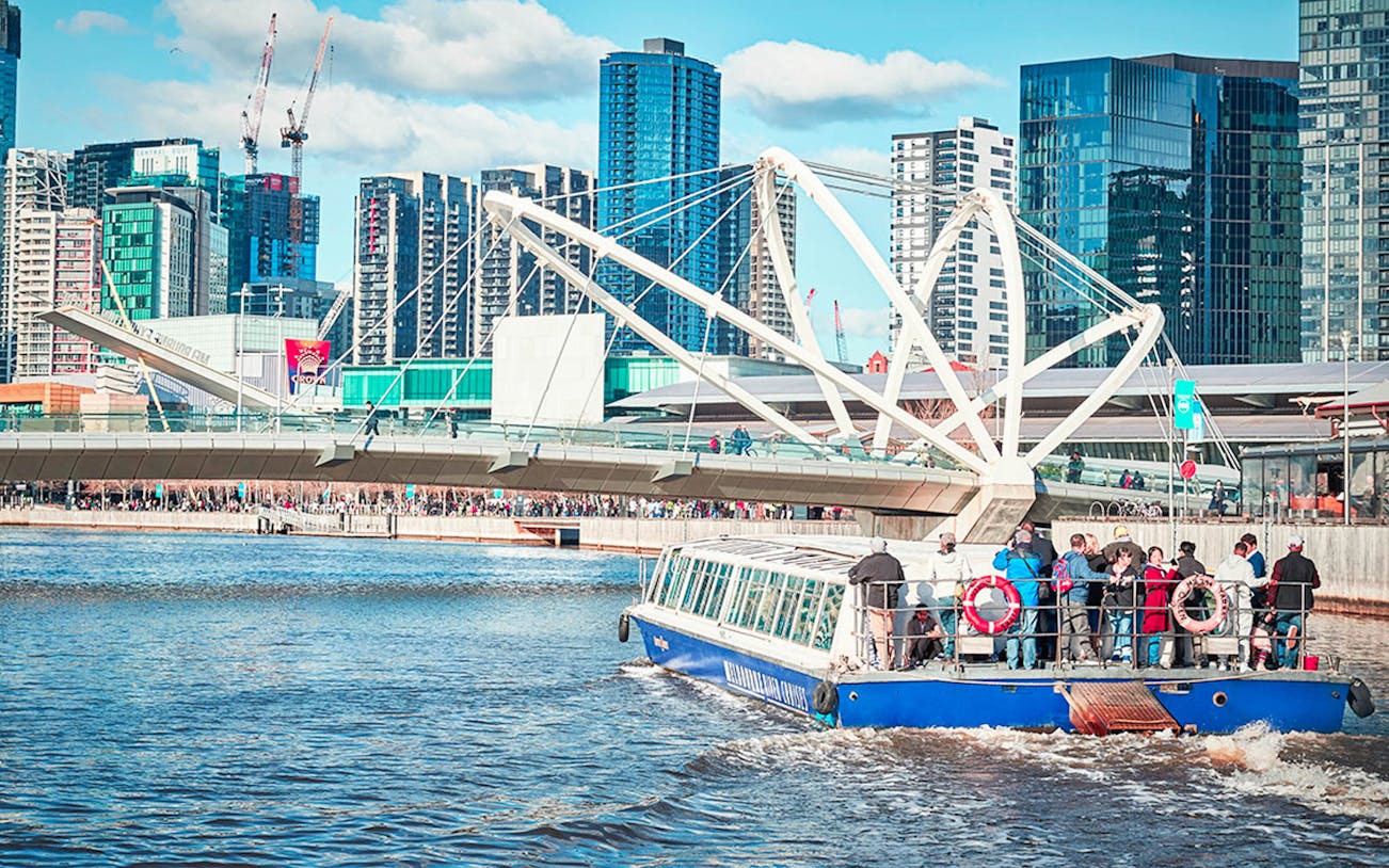 Tourists on a river cruise boat passing under a modern bridge in a cityscape.