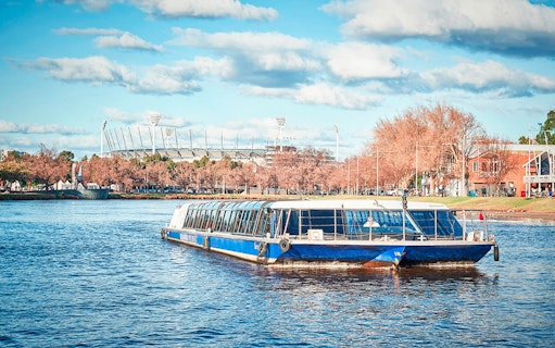River cruise boat on a scenic waterway with a stadium and trees in the background.
