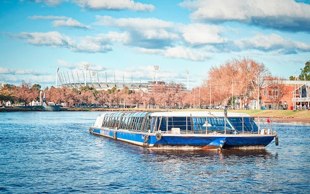 River cruise boat on a scenic waterway with a stadium and trees in the background.