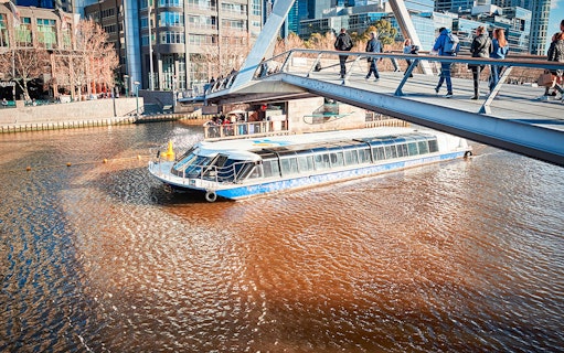 River cruise boat passing under a pedestrian bridge in a cityscape setting.