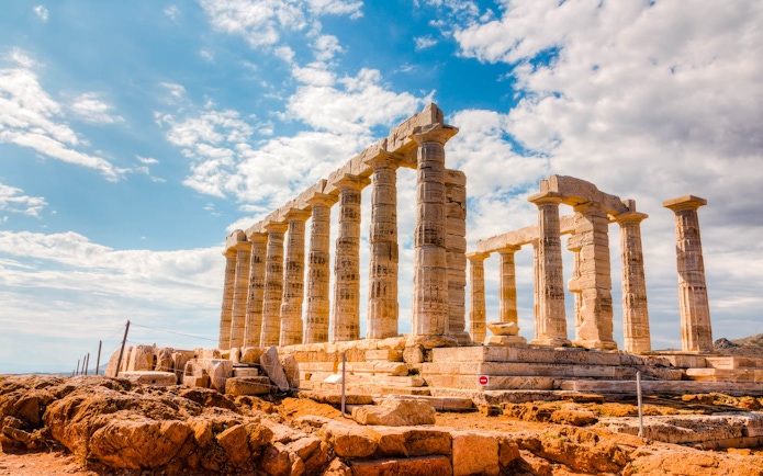 Temple of Poseidon ruins at Cape Sounion under a blue sky, viewed from rocky terrain.