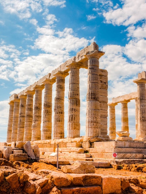 Temple of Poseidon ruins at Cape Sounion under a blue sky, viewed from rocky terrain.