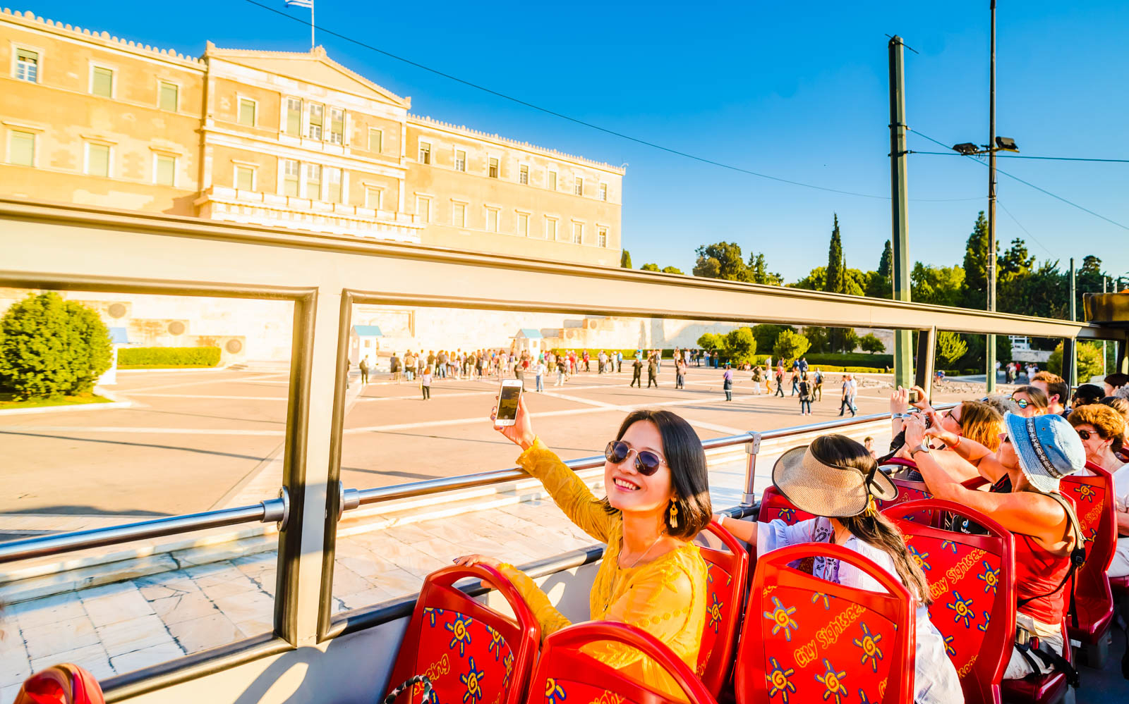 Tourists on an open-top bus in Athens, viewing the Hellenic Parliament building.