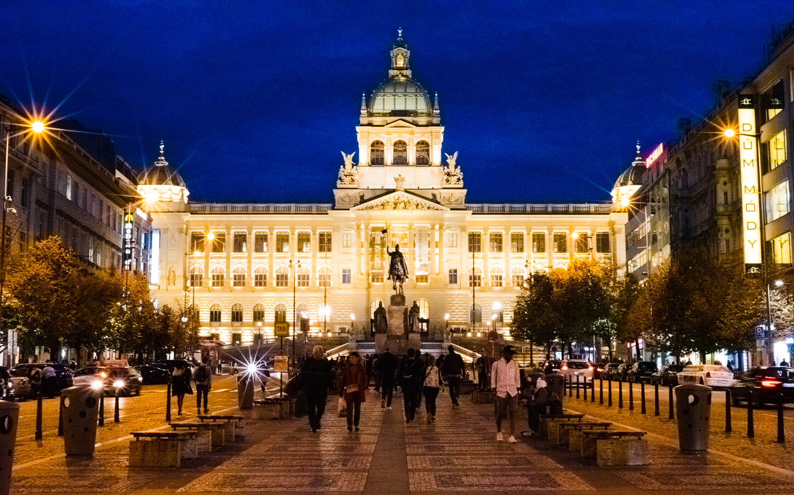 Prague National Museum illuminated at night with people walking in front.