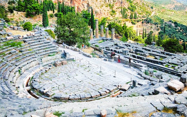 Ancient theater ruins at Delphi, Greece, surrounded by lush greenery and distant mountains.