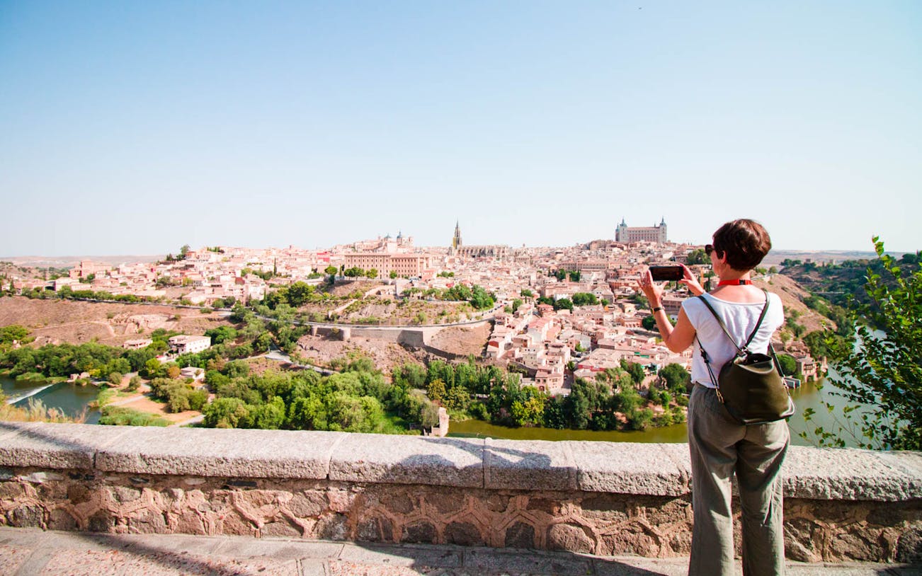 Person photographing Toledo cityscape from a viewpoint during guided tour.