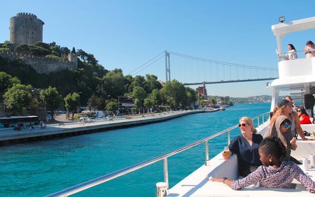 Bosphorus cruise with tourists enjoying lunch, Rumeli Fortress, and bridge in view.