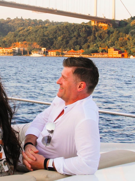 Couple enjoying Bosphorus sunset yacht cruise with drinks, Istanbul bridge in background.