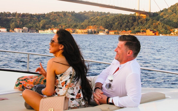 Couple enjoying Bosphorus sunset yacht cruise with drinks, Istanbul bridge in background.