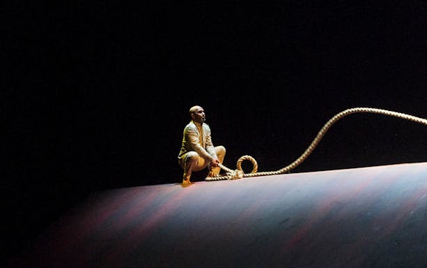 Performer crouching on stage with rope in Akram Khan Company’s XENOS.