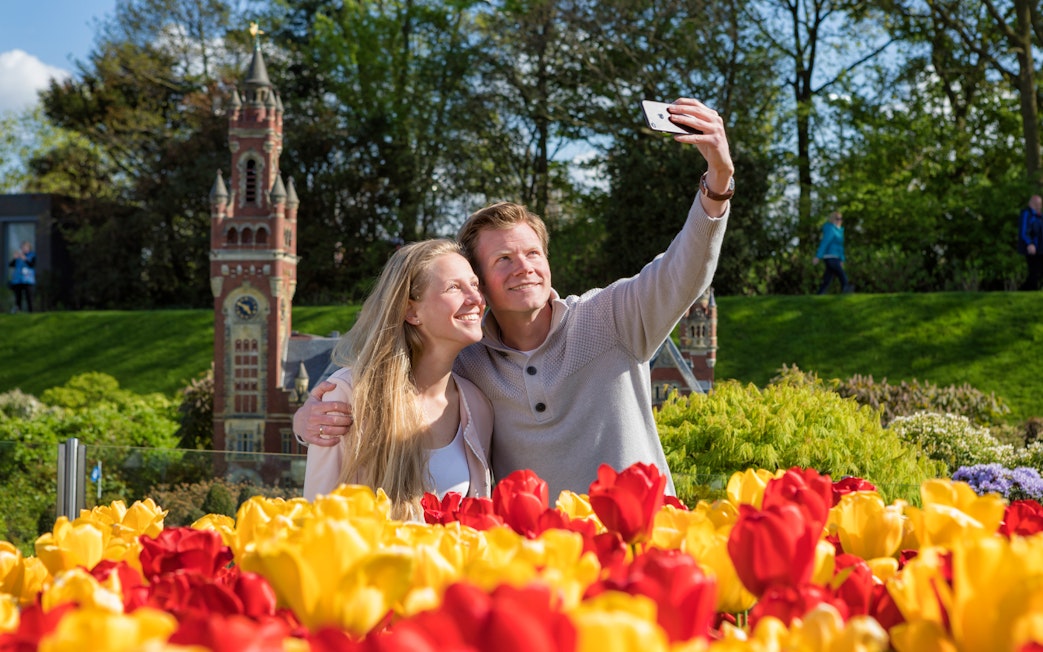 Couple taking selfie at Madurodam Miniature Park with tulips and clock tower in view.