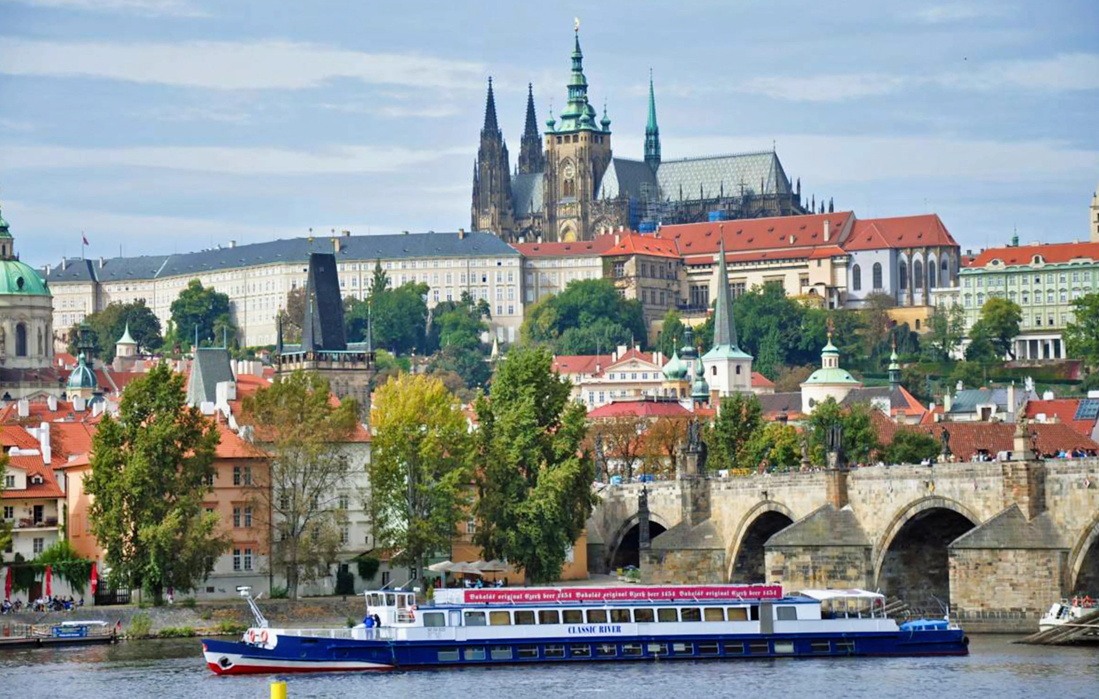 Panoramic view of Prague Castle and Charles Bridge with a cruise boat on the Vltava River.