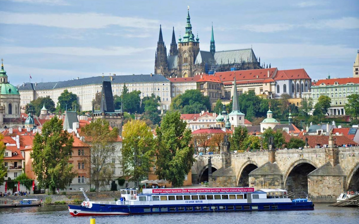Panoramic view of Prague Castle and Charles Bridge with a cruise boat on the Vltava River.