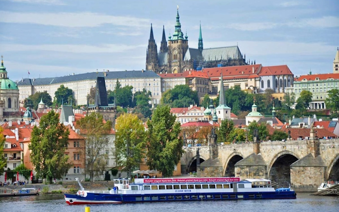 Panoramic view of Prague Castle and Charles Bridge with a cruise boat on the Vltava River.